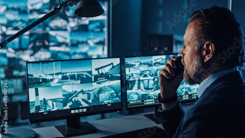 Male Officer Works on a Computer with Surveillance CCTV Video in a Harbour Monitoring Center with Multiple Cameras on a Big Digital Screen. Employee Uses Radio to Give an Order or Report.