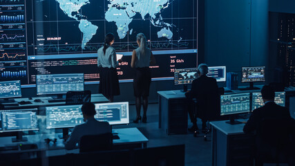 Poster - Two Software Engineers Talking Next to a Big Screen in a Modern Monitoring Office with Live Analysis Feed with Charts. Monitoring Room Big Data Scientists and Managers Sit in Front of Computers.
