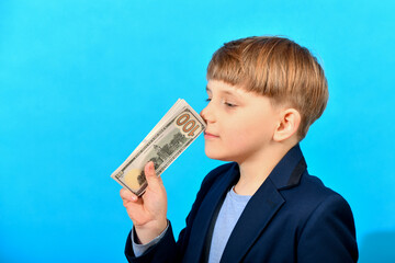 Boy in a suit sniffing dollar bills on a blue background. Greed for wealth concept.