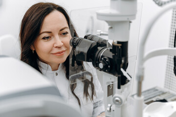 Young beautiful woman doctor / eye specialist / optometrist in an ophthalmologic clinic examines a patient with microscopy.