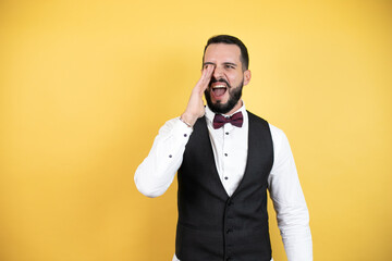 Wall Mural - Young man with beard wearing bow tie and vest shouting and screaming loud to side with hands on mouth