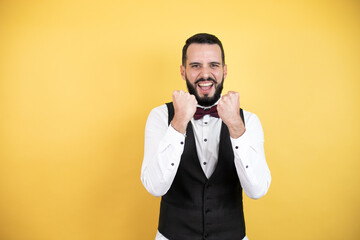 Wall Mural - Young man with beard wearing bow tie and vest very happy and excited making winner gesture with raised arms, smiling and screaming for success.