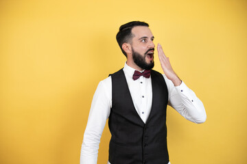 Wall Mural - Young man with beard wearing bow tie and vest covering mouth with hand, shocked and afraid for mistake. surprised expression