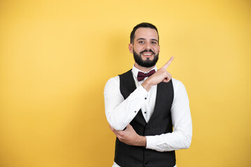 Wall Mural - Young man with beard wearing bow tie and vest amazed and pointing with hand and finger to the side