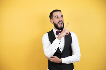 Wall Mural - Young man with beard wearing bow tie and vest amazed and pointing with hand and finger to the side