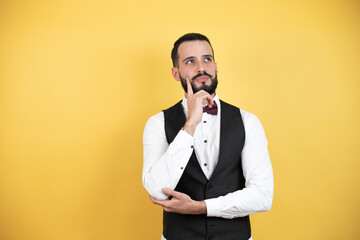 Wall Mural - Young man with beard wearing bow tie and vest serious face thinking about question with hand on chin, thoughtful about confusing idea