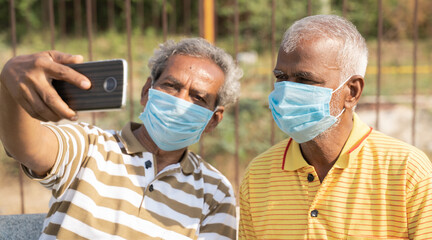 elderly friends with medical face mask taking selfie while sitting at park - concept of happy senior people friendship and meetup after lockdown due to coronavirus covid-19 pandemic.