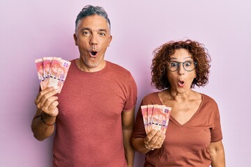 Sticker - Beautiful middle age couple holding south african 50 rand banknotes scared and amazed with open mouth for surprise, disbelief face