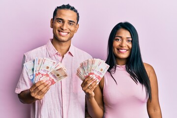 Wall Mural - Young latin couple holding colombian pesos looking positive and happy standing and smiling with a confident smile showing teeth