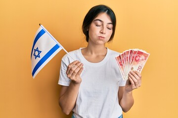 Wall Mural - Young caucasian woman holding israel flag and shekels banknotes puffing cheeks with funny face. mouth inflated with air, catching air.