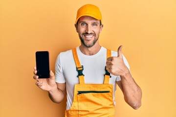 Wall Mural - Young handsome man wearing handyman uniform showing smartphone screen smiling happy and positive, thumb up doing excellent and approval sign