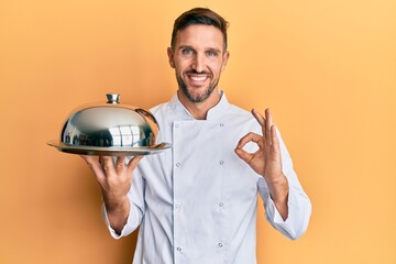 Wall Mural - Handsome man with beard wearing chef uniform holding silver tray doing ok sign with fingers, smiling friendly gesturing excellent symbol