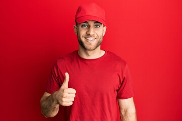 Young caucasian man wearing delivery uniform and cap smiling happy and positive, thumb up doing excellent and approval sign