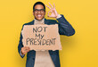 © Krakenimages.com - Young handsome hispanic man holding not my president protest banner doing ok sign with fingers, smiling friendly gesturing excellent symbol