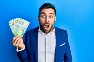 Canvas Print - Young hispanic businessman wearing business suit holding russian ruble banknotes scared and amazed with open mouth for surprise, disbelief face