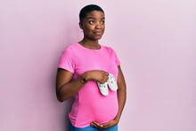 Young African American Woman Expecting A Baby Holding Shoes Smiling Looking To The Side And Staring Away Thinking.