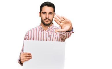 Young hispanic man holding blank empty banner with open hand doing stop sign with serious and confident expression, defense gesture