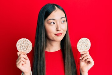 Poster - Young chinese woman eating healthy rice crackers smiling looking to the side and staring away thinking.