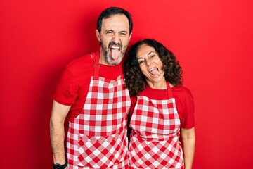 Wall Mural - Middle age couple of hispanic woman and man wearing cook apron sticking tongue out happy with funny expression. emotion concept.