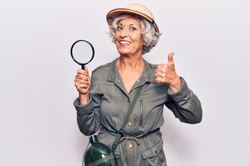 Senior grey-haired woman wearing explorer hat holding magnifying glass smiling happy and positive, thumb up doing excellent and approval sign