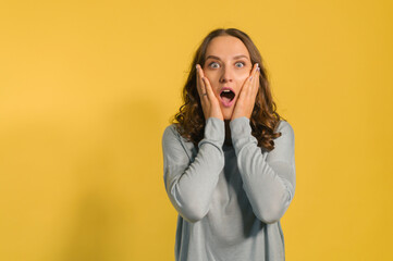 Shocked and amazed young attractive woman looks at the camera isolated on yellow, a curly caucasian girl screams surprised, holding cheeks, incredible sales or deal