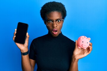 Sticker - Young african american girl holding piggy bank and smartphone afraid and shocked with surprise and amazed expression, fear and excited face.