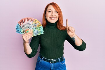 Canvas Print - Beautiful redhead woman holding australian dollars smiling with an idea or question pointing finger with happy face, number one