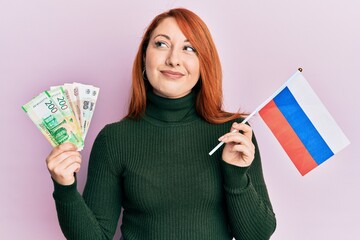 Poster - Beautiful redhead woman holding russian ruble banknotes and russia flag smiling looking to the side and staring away thinking.