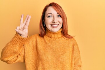 Canvas Print - Beautiful redhead woman wearing casual winter sweater over yellow background showing and pointing up with fingers number three while smiling confident and happy.