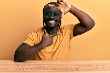 Poster - Handsome young black man wearing casual clothes sitting on the table smiling making frame with hands and fingers with happy face. creativity and photography concept.