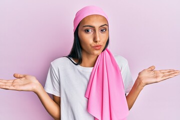 Poster - Young brunette woman wearing breast cancer support pink scarf clueless and confused expression with arms and hands raised. doubt concept.