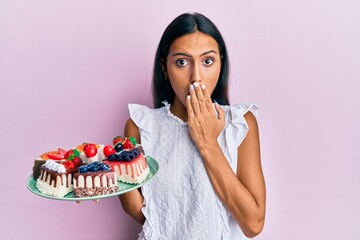 Wall Mural - Young brunette woman holding cake slices covering mouth with hand, shocked and afraid for mistake. surprised expression