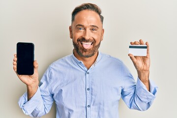 Sticker - Handsome middle age man holding smartphone and credit card sticking tongue out happy with funny expression.