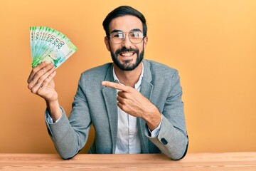 Young hispanic man holding russian ruble banknotes sitting on the table smiling happy pointing with hand and finger