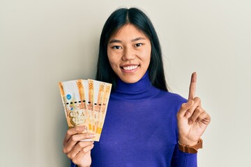 Sticker - Young chinese woman holding 500 philippine peso banknotes smiling with an idea or question pointing finger with happy face, number one