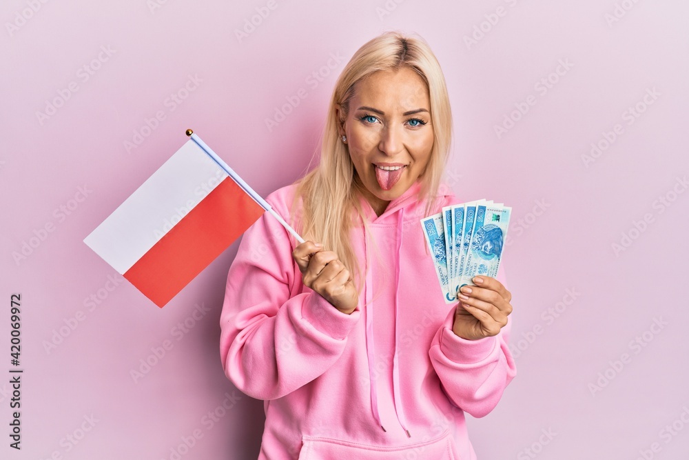 Young blonde woman holding poland flag and zloty banknotes sticking tongue out happy with funny expression. - obrazy, fototapety, plakaty 