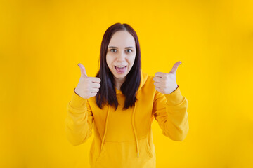 Wall Mural - Portrait of young woman smiling and showing thumbs up on yellow background. Close up of happy brunette in yellow hoodie showing gesture of approval.