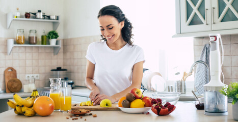 cute beautiful and happy young brunette woman in the kitchen at home is preparing fruit vegan salad 