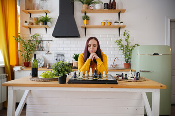 Young woman playing chess on kitchen table. Female plays in logical board game with herself in kitchen.