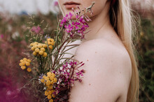 Young Woman With Naked Sholders Holding Bouquet Of Yellow And Purple Wild Flowers Standing In Green Field At Summer. Caucasian Girl With Blonde Hair. Outdoors Portrait In Countryside.