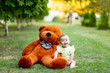 © Any Grant - a smiling little girl is sitting on the green grass with a large Teddy bear in a yellow summer dress in summer