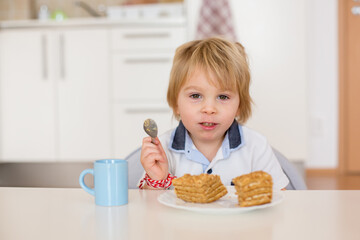 Wall Mural - Cute preschool boy, blond child, eating piece of cake and drinking milk at home