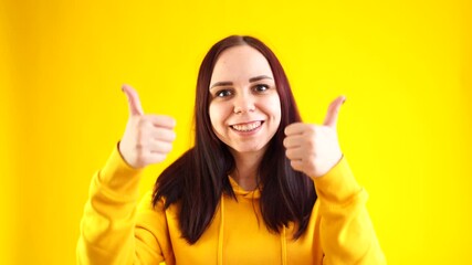 Wall Mural - Portrait of young woman smiling and showing thumbs up on yellow background. Close up of happy brunette in yellow hoodie showing gesture of approval.