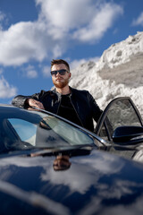 Wall Mural - Attractive man standing near opened car door. Nice view of mountain and sky. Stylish combination of black and white.