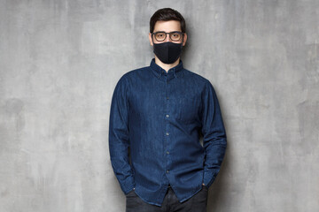 Poster - Young man wearing denim shirt, glasses and black mask, standing against gray textured wall