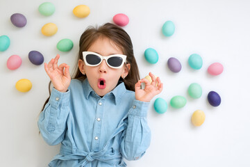 A girl in sunglasses makes a gesture with her hands. Baby on a white background with Easter eggs. The child opened his mouth in surprise