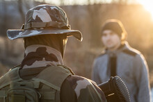 Back View Of Unknown Armed Soldier Standing Against A Migrant Or Terrorist Or Criminal In Sunny Evening In Nature During Border Patrol Or Mission