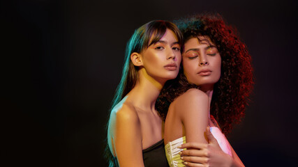 Wall Mural - Two stylish young women with professional art makeup posing together in neon light isolated over black background