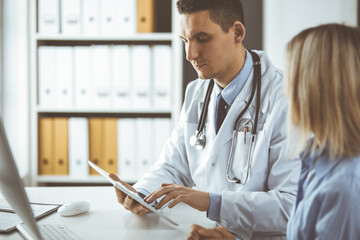 Unknown male doctor and patient woman discussing current health examination while sitting in clinic and using tablet computer. Perfect medical service in hospital. Medicine and healthcare concept