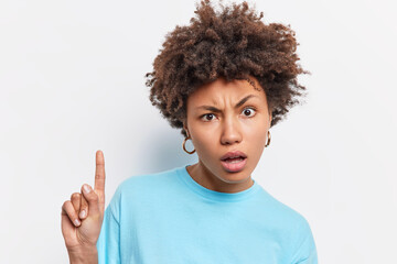 Wall Mural - Headshot of displeased curly haired Afro American woman looks puzzled at camera indicates index finger above shows something amazing dressed in casula blue t shirt isolated over white background